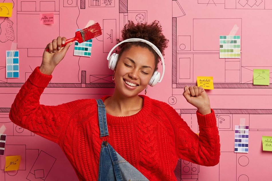 Amused relaxed woman dances happily, raises arms with paint brush, listens music in wireless headphones as does house repair, closes eyes with pleasure, moves against modern house design project